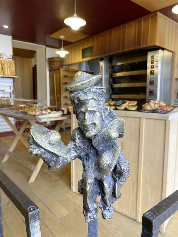 L’intérieur de la boutique : le fournil ouvert, son four à soles, les planches de viennoiseries, et la statue de bronze d’un boulanger portant deux pains.