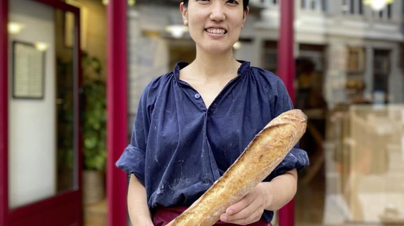 Mei Narusawa devant la porte bordeaux de sa boulangerie, une baguette de tradition dans les bras.