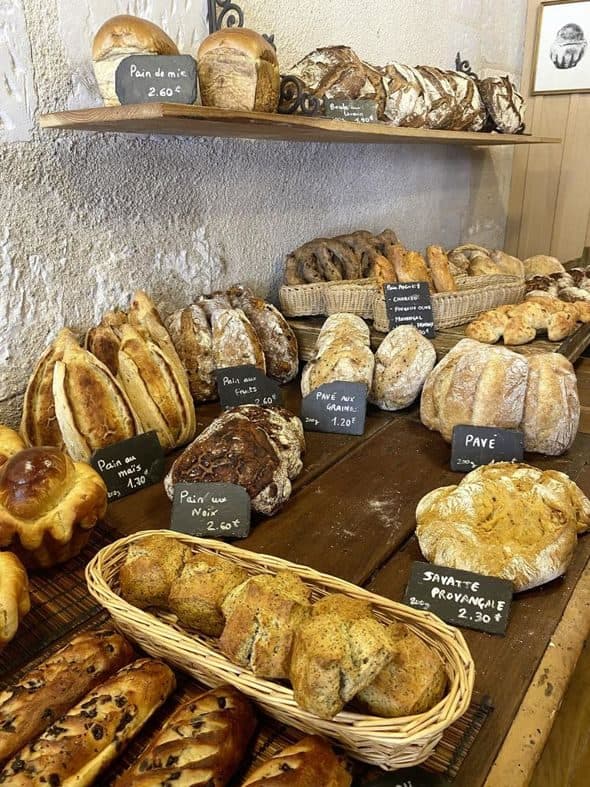 Les pains du jour posés sur les planches de bois de la boutique : boules au levain, pavés aux graines, pain aux noix, savatte provençale, avec leurs ardoises manuscrites.